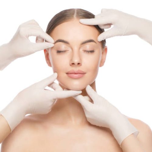 Close-up portrait of young woman standing with naked shoulders and neck, face is touched by beauticians in gloves, preparing her for plastic surgery procedures, isolated on white background